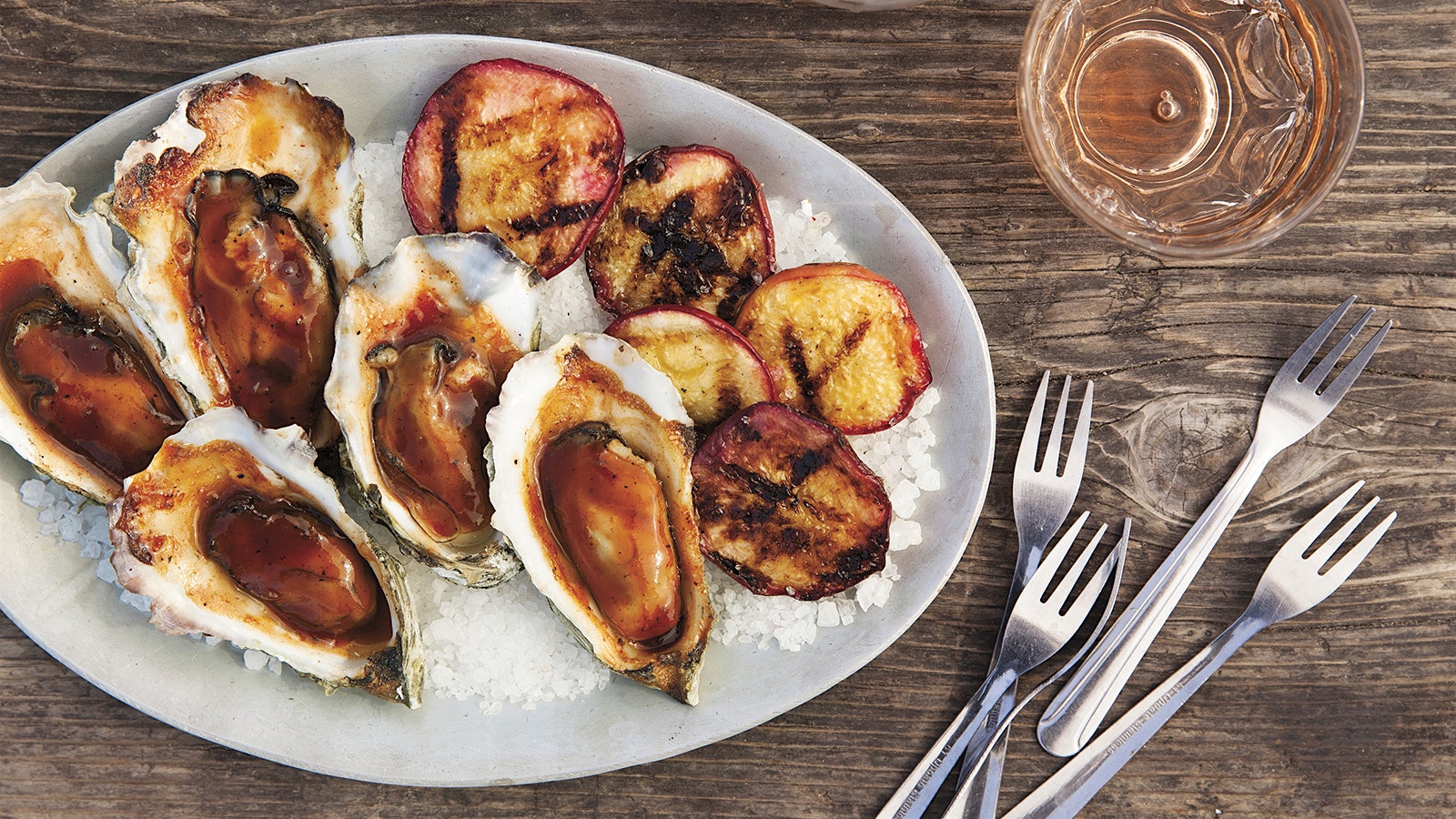  A plate of grilled oysters, topped with peach BBQ sauce, on crushed ice, accompanied by roasted summer peaches and glasses of rosé 